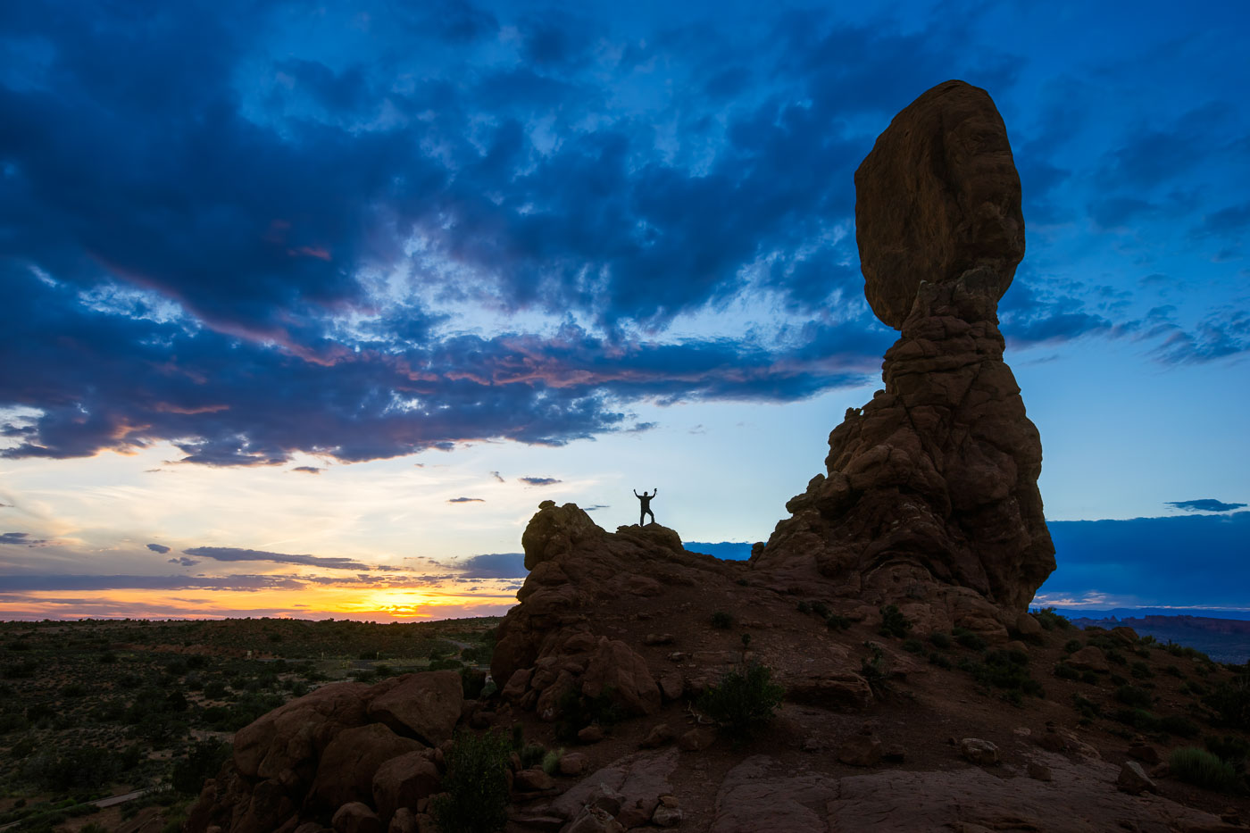 Balanced Rock silhouette at sunset with person on nearby rock, Arches National Park